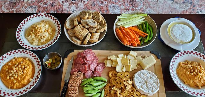 A table with various bowls of hummus, cut vegetables and bread, in the middle is a cutting board with meats and cheese.