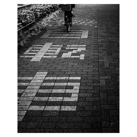 A textured sidewalk in monochrome features the kanji characters for “bicycle” (自転車) embedded into the brickwork in a charmingly pixelated style. The chunky, blocky font recalls retro gaming graphics, a lo-fi declaration of cycling territory. A rider approaches in the background, front wheel perfectly aligned with the path's utilitarian intent.