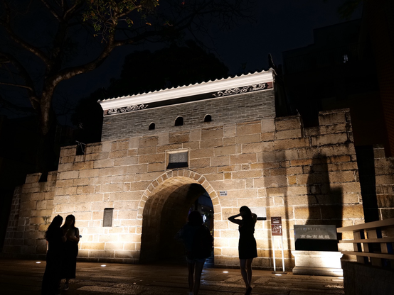 An old Chinese gate is illuminated at night. A sign to the side says that it is Nantou Ancient Town, which is in Shenzhen. Some silhouetted people in front are taking photos 