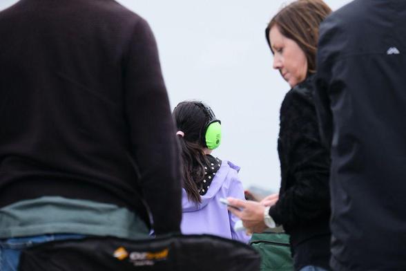 Photo of a young girl in a purple jacket wearing large, yellow green protection earmuffs while watching the flying display at an airshow. 
