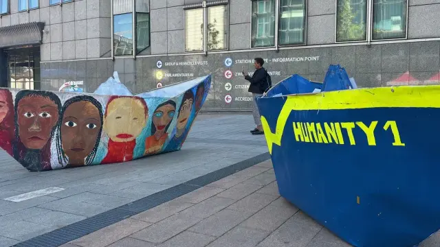 Das Bild zeigt eine städtische Szene mit künstlerisch gestalteten Objekten in Form von Booten. Es gibt zwei große Bootsstrukturen auf einem gepflasterten Boden vor einem modernen Gebäude. 

1. **Linkes Boot**: Es ist mit Gesichtern von Menschen in verschiedenen Stilen und Farben bemalt, die Vielfalt und Menschlichkeit darstellen könnten. Die Gesichter sind frontal dargestellt und scheinen freundlich und ausdrucksstark.

2. **Rechtes Boot**: Auf diesem Boot steht der Text "HUMANITY 1" in gelber Schrift auf blauem Hintergrund, was möglicherweise eine Botschaft der Einheit oder Solidarität vermitteln soll.

3. **Umgebung**: Im Hintergrund ist ein modernes Gebäude mit großen Fenstern und einer Wand zu sehen. Einige Schilder und ein Mensch, der an den Booten vorbeigeht, sind ebenfalls sichtbar.

Die Szene wirkt wie eine künstlerische Installation oder ein öffentlicher Kunstbeitrag, der auf gesellschaftliche Themen aufmerksam machen könnte.
