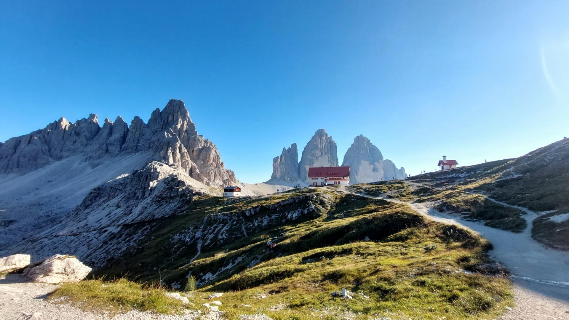 Berglandschaft mit den Drei Zinnen zentral im Hintergrund. Davor die Dreizinnenhütte. Rechts im Bild der Wanderweg zur Hütte, der durch Wiesengelände führt. Ein sonniger, wolkenloser Tag. Berglandschaft mit den Drei Zinnen zentral im Hintergrund. Davor die Dreizinnenhütte. Rechts im Bild der Wanderweg zur Hütte, der durch Wiesengelände führt. Ein sonniger, wolkenloser Tag.