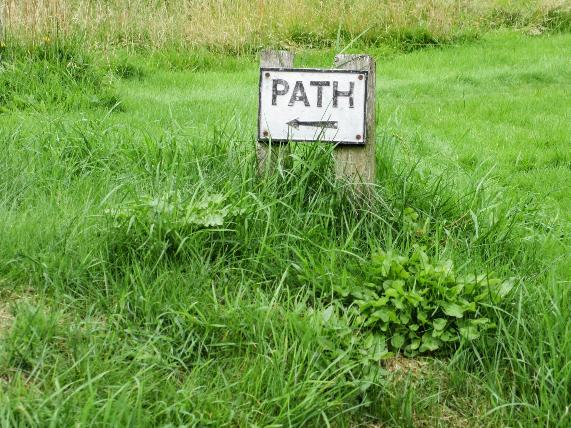 Ein kleines weisses Blechschild mit schwarzer Beschriftung "Path" und einem Pfeil nach links. Das Schild steht auf einer Wiese. Es ist an zwei kurze, etwa 30 Zentimeter hohe, Holzlatten genagelt. Ein kleines weisses Blechschild mit schwarzer Beschriftung "Path" und einem Pfeil nach links. Das Schild steht auf einer Wiese. Es ist an zwei kurze, etwa 30 Zentimeter hohe, Holzlatten genagelt.