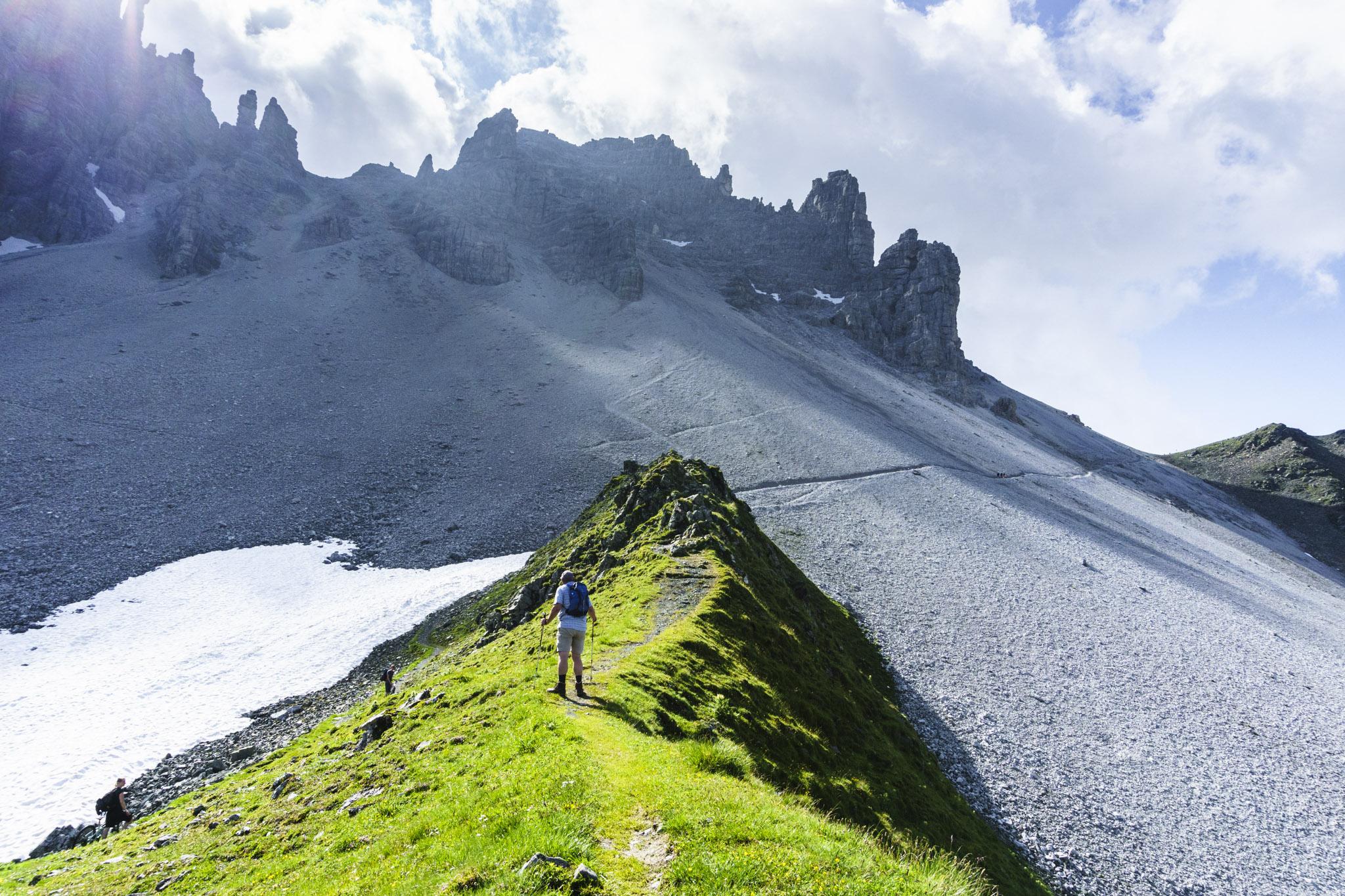 Ein Wanderweg, der auf einem kleinen Wiesengrat verläuft. Drum herum Schottergelände. Im Hintergrund setzt sich der Weg leicht ansteigend durch den Schotter fort. Im Bildhintergrund links Felsberge. Ein sonniger, aber wolkenreicher Sommertag. Ein Wanderweg, der auf einem kleinen Wiesengrat verläuft. Drum herum Schottergelände. Im Hintergrund setzt sich der Weg leicht ansteigend durch den Schotter fort. Im Bildhintergrund links Felsberge. Ein sonniger, aber wolkenreicher Sommertag.
