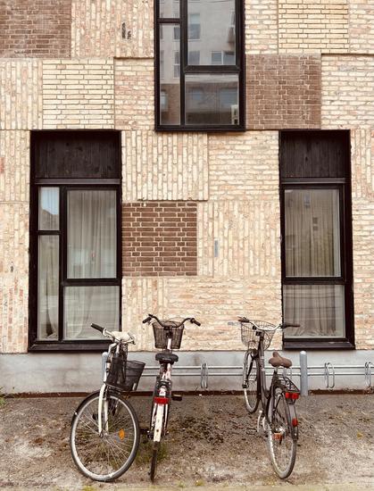 A building facade with a unique patchwork of brick textures and colors features multiple windows. In the foreground, three bicycles are parked, two of which have baskets, against a concrete surface.