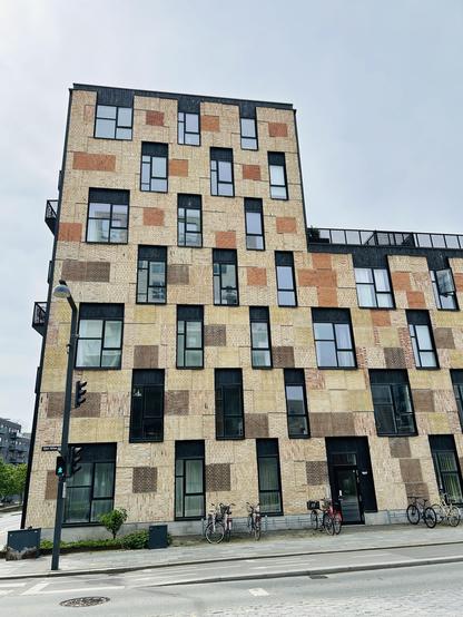 A modern building featuring a unique facade with a patchwork of different brick colors and window designs. Bicycles are parked along the front, and traffic lights are visible on the street. The sky is partly cloudy.