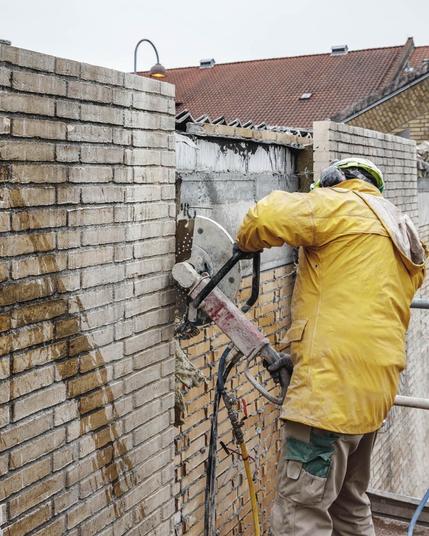 A construction worker in a yellow jacket operates a concrete cutting saw on a brick wall. The scene depicts a demolition or renovation project, with debris and materials visible around the work area. District rooftops can be seen in the background.