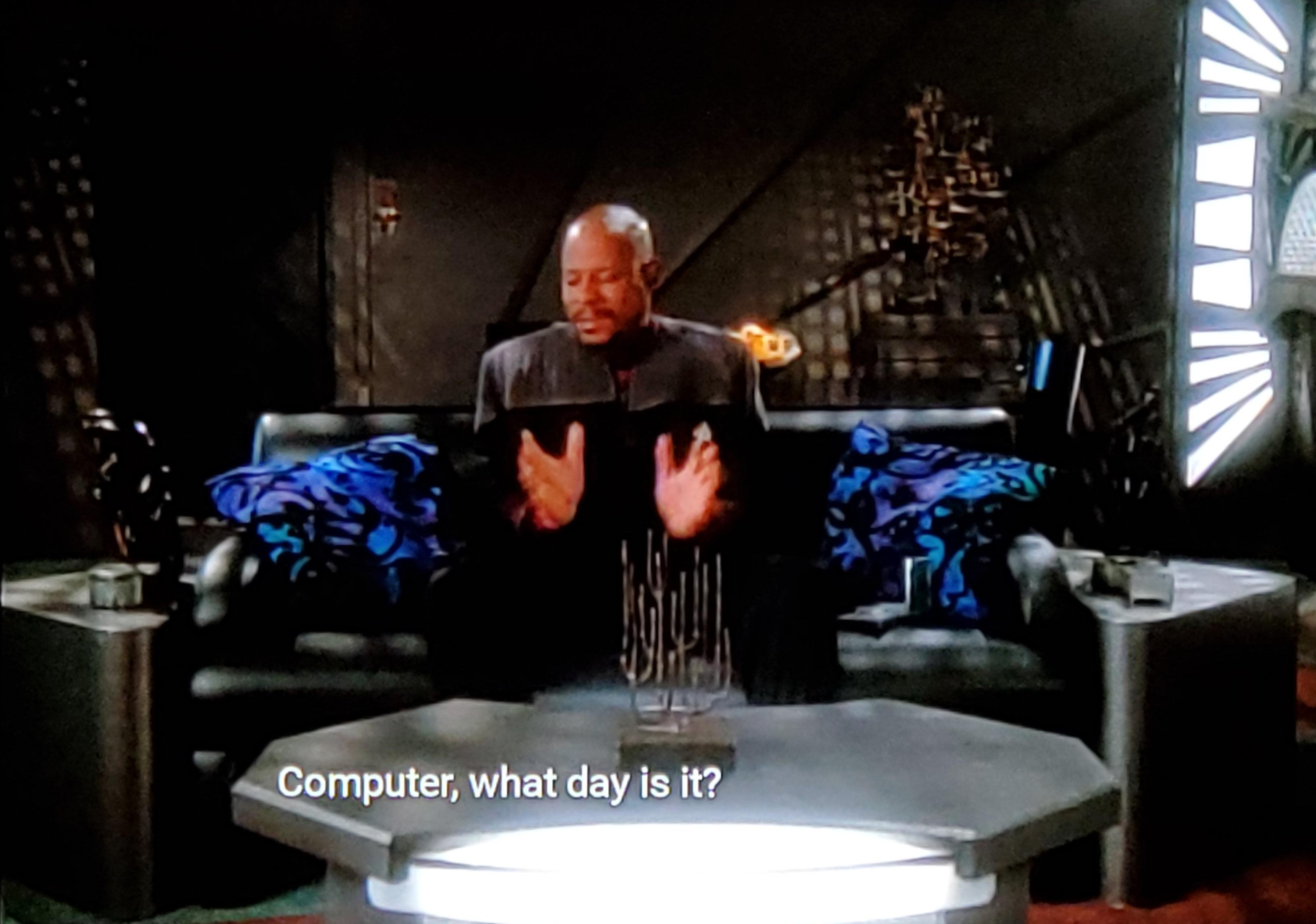 Star Trek Deep Space Nine scene. The Sisko sits in quarters on a couch behind a coffee table. There's some very blue and purple pillows on the couch, in an otherwise very dimly lit room. He's holding his hands up as if he's animatedly speaking. Closed caption reads, "Computer, what day is it?"