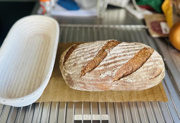 Ein länglich geformtes Brot mit drei kräftigen diagonalen Einschnitten liegt auf Backpapier und einem Gitter zum Auskühlen. Daneben noch das verwendete Gärkörbchen.