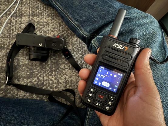 An hand holds a handheld network radio with digital display, labeled "KSUN," while a camera is placed beside them on a carpet.