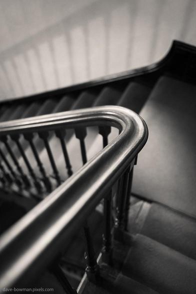 A photograph of the staircase at Blickling House, taken from above, where the curve of the bannister leads the eye downward in a smooth arc. Shadows cast by the evenly spaced balusters fall gently across the adjacent wall, creating a quiet, rhythmic pattern. The monochrome tones highlight the textures of the wood and stone, subtly reflecting the historic character of this Jacobean mansion in Norfolk.