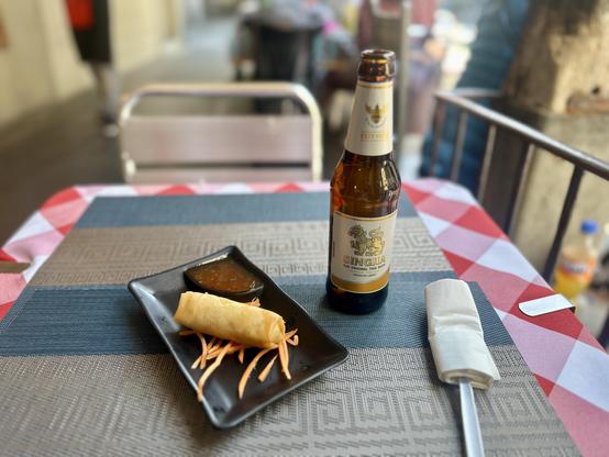Eine Frühlingsrolle auf Deko (Karottenstreifen) auf einem rechteckigen Teller mit etwas süss-scharfer Sauce. Dahinter eine Flasche Singha Bier.
