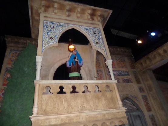 n the #IslamicEmpire scene, on the right, four men sit around a table on pillows on the floor discussing topics. One man has two books right next to him and another has a wooden book holder that holds the book open to a speci
