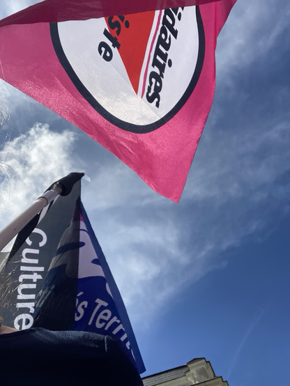 Photo en contre plongée, 3 drapeaux flottent dans le vent devant un ciel bleu et nuageux : le drapeau rose avec un triangle rouge au milieu pour Solidaires Antifasciste, le drapeau noir aux écritures blanches de SUD Culture Solidaires et le drapeau bleu foncé de SUD Collectivités Territoriales