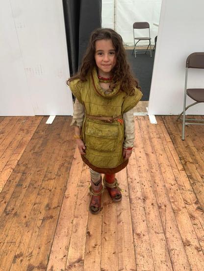 The actress is wearing her clothes from the show. She is in a room with a wooden floor, we can see two chairs on the right hand side.