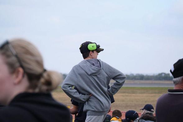 Photo of a young boy in a grey hoodie wearing large, green hearing protection earmuffs above his ears at an airshow. 