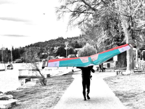 Schwarzweissfotografie mit Selektivfarben einer Person von hinten, die einen Wingsurf-Flügel auf einem Gehweg trägt. 