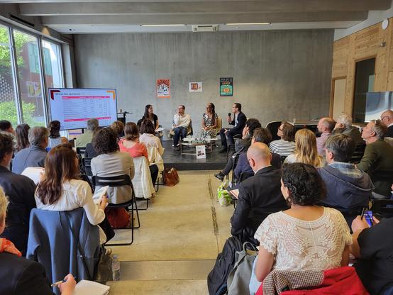 Un groupe d'adultes assis face à une scène où quatre adultes parlent au micro