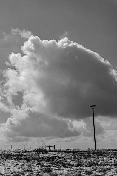 Black and white image of a snow-dusted field under a large, dramatic cloud. A single raptor perch pole stands to the right, silhouetted against the sky, with a fence and a second perch in the background.