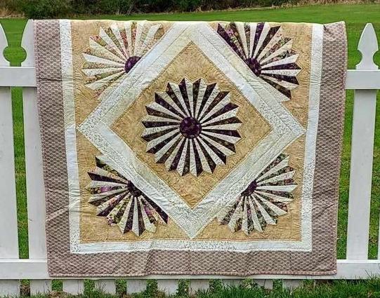 a  quilt, it has a tan checkered border, inside is a diamond shape, in the center of the diamond is a sharp wheel of light tan and darker fabrics, on each line of the diamond is a half-wheel 