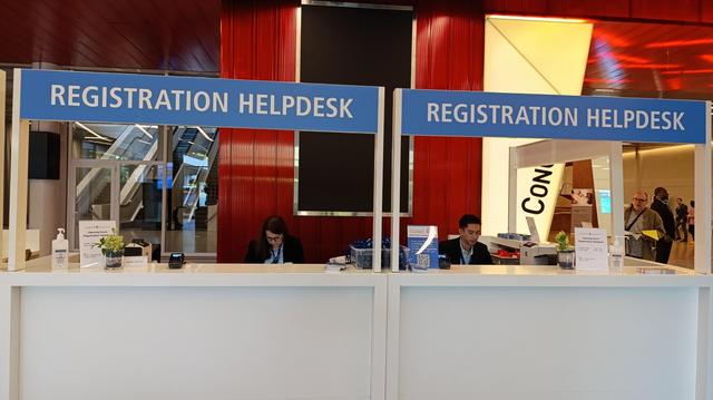 Two Congrex Switzerland staff members are seated behind a sleek white helpdesk marked “Registration Helpdesk” with bright blue signage. They are focused on their laptops, ready to assist participants. Behind them is a vibrant red wall and the entrance area of the event venue.