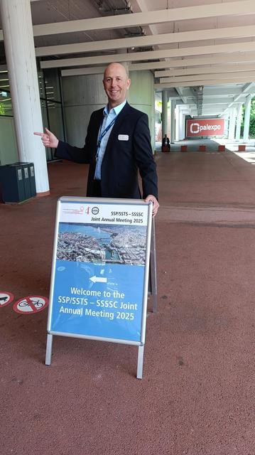 A Congrex Switzerland team member in formal attire stands outdoors next to a freestanding sign for the SSP/SSTS – SSSSC Joint Annual Meeting 2025. He smiles and gestures towards the sign, which displays a photo of Geneva and the event title. The location is just outside the Palexpo venue, under a modern concrete overhang.
