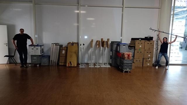Two team members stand in a storage room surrounded by packed boxes, signage stands, crates, and technical equipment. One person poses playfully holding a metal coat rack like a victory trophy, while the other stands casually beside stacked gear. It’s a candid behind-the-scenes glimpse.