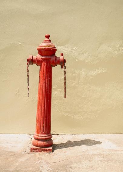 A photograph of a red Penang fire hydrant standing in sunlight against a light olive green wall, its vivid colour creating a strong contrast with the muted background. Light and shadow fall across the scene, adding warmth and depth to the straightforward composition.