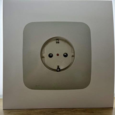 Eine Steckdose, die auf einer weißen Wandplatte montiert ist. Die Steckdose hat zwei runde Löcher und einen kleinen roten Punkt in der Mitte.