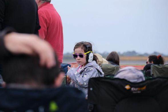 Photo of a young girl in a pale blue jacket wearing large, white hearing protection earmuffs at an airshow.