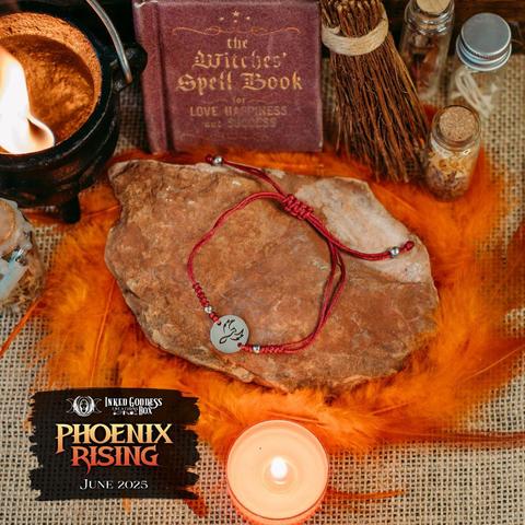 A bracelet made of red knotted cord sits on a flat red rock, surrounded by red and orange feathers. The end is adjustable with slipknots accented with small silver beads. The focal element is a silver coin charm with an abstract phoenix stamped into it. There are knots in the cord to either side accented with small silver beads. This is one of the items in the exclusive June 2025 "Phoenix Rising" Inked Goddess Creations Box.