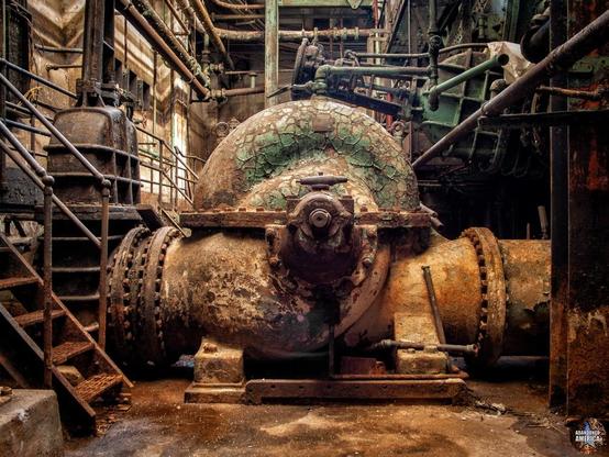 A large water valve (?) that resembles a seashell covered in rust and peeling paint is in the foreground. A short stairway to another platform is to the left. I don't know what the other machinery is tbh