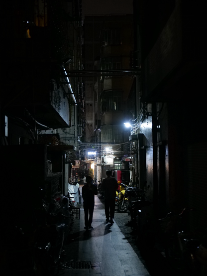 Two people walk down a dark alley. Some lights on either side illuminate their path a little. Surrounding them are buildings and cables 