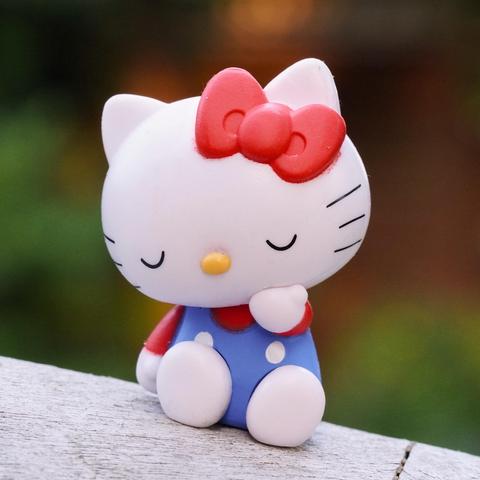 A small figurine of the Hello Kitty character, with a red bow, sitting with eyes closed. The cat has a white face and wears a blue outfit with red sleeves and white polka dots. The background is blurred with green and warm tones.