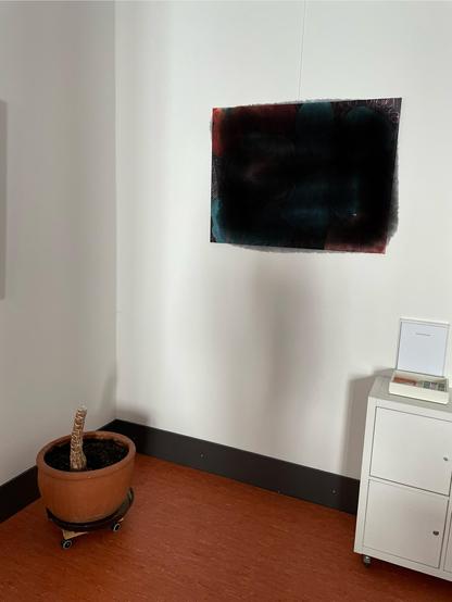 Das Bild zeigt einen Raum mit einem minimalistischen Design. An der weißen Wand hängt ein abstraktes Gemälde in dunklen Farben, hauptsächlich Schwarz, Blau und Rot, mit einer unregelmäßigen, organischen Form. Unterhalb des Gemäldes befindet sich ein kleiner, weißer Schrank mit zwei Schubladen, auf dem ein kleines, offenes Buch oder ein Kartenständer steht. In der linken Ecke des Raumes steht ein Terrakotta-Topf mit einem angeschnittenen Baumstamm, der auf einem Rollwagen steht. Der Boden ist aus Plastik und hat einen warmen, braunen Farbton. Die Wände sind weiß, und die Unterseite der Wände ist mit einem schwarzen Abschluss versehen.