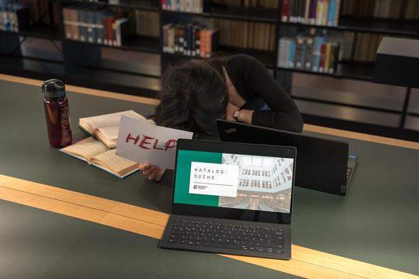 Deutsch: Im Hintergrund ist eine Studentin zu sehen, die an einem Arbeitsplatz der Universitätsbibliothek Chemnitz sitzt und ihren Kopf verzweifelt auf der Tischplatte gebettet hat. Vor sich hat sie einen Laptop, einige aufgeschlagene Bücher und eine Wasserflasche stehen - in der Hand hält sie ein Schild mit der Aufschrift "HELP" (dt. Hilfe). Im Vordergrund ist ein weiterer Laptop zu sehen, auf dessen Bildschirm eine Präsentationsseite mit dem Titel "Katalogsuche" geöffnet ist. // English: A student can be seen in the background, sitting at a desk in the Chemnitz University Library with her head resting desperately on the tabletop. In front of her is a laptop, some open books and a bottle of water - in her hand she is holding a sign that reads ‘HELP’. Another laptop can be seen in the foreground, with a presentation page entitled ‘Catalogue search’ open on its screen.