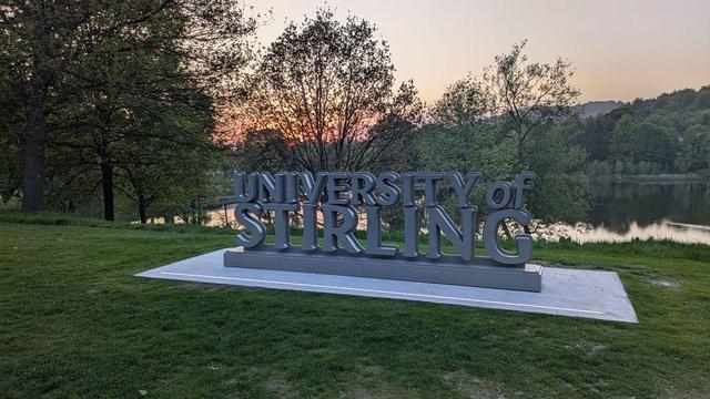 A statue of the University of Stirling logo, standing next to a lake, with mountains, trees and the sunset in the background.