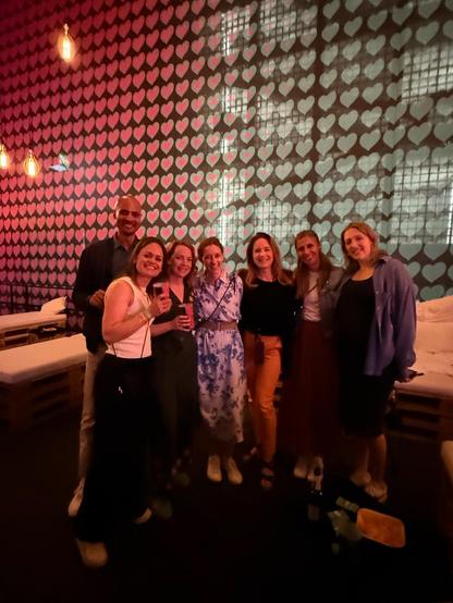 A group of smiling Congrex Switzerland team members pose for a photo at the Eurovision VIP Lounge. They’re standing in front of a wall covered in a pattern of glowing pink hearts. The atmosphere is relaxed and celebratory, with warm ambient lighting and casual seating in the background.