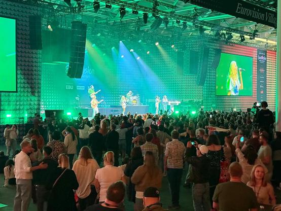 A lively crowd watches a Eurovision concert on stage inside the Eurovision Village. Green stage lights wash over the audience as a band performs under the iconic heart-covered backdrop. A large screen to the right shows a close-up of the performer. The venue is packed and buzzing with excitement.