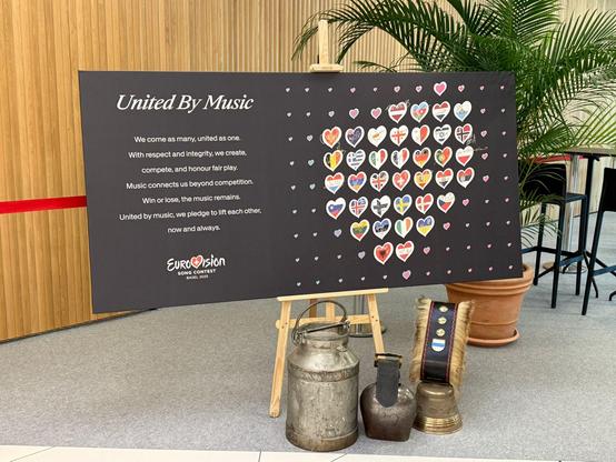 A black board on an easel with the Eurovision Song Contest 2025 branding. It displays a unifying message titled "United By Music", surrounded by colourful heart-shaped flags of participating countries. Decorative props including cowbells and old milk jugs are placed beneath the board, adding a local Swiss touch.