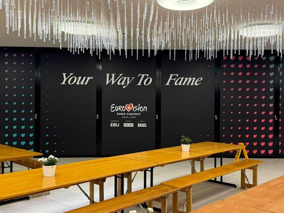 A stylish black wall decorated with pink and teal heart patterns and the slogan "Your Way To Fame" in large white letters. Below it, the Eurovision Song Contest Basel 2025 logo appears alongside organiser logos (EBU, SRG SSR, Basel). In front, wooden benches and tables are neatly arranged under an elegant ceiling of suspended LED rods.
