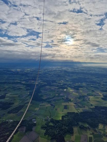 A landscape of fields and woods, taken from a hot air balloon, of which you can see a cable in the picture. The sky is cloudy but shows shafts of sunlight.