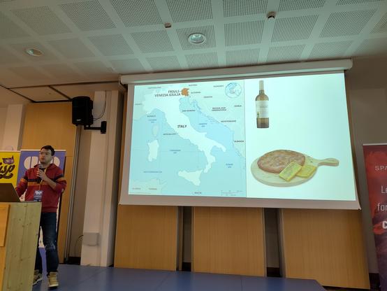 Luigi standing in front of a projector slide showing Friuli on the italian map, together with wine and frico, both traditional of the region.