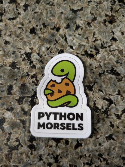 A sticker of an adorable green snake hugging a vegan chocolate chip cookie with the text "Python Morsels" below it.