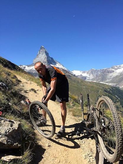 Auf hohem Berg im Gebirge steht ein Mann und pumpt sein ausgebautes Vorderrad auf. Das Rest-Fahrrad steht daneben auf dem Kopf gestellt auf einem schmalem Gebirgspfad. Im Hintergrund sind hohe Berggipfel vor blauem, wolkenlosen Himmel zu sehen.