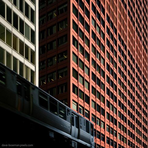 A photograph of a Chicago L train passing between tall office blocks, captured from a low angle. The train appears slightly blurred, suggesting movement, while the background features sharp, repetitive patterns of red and white buildings. The contrast between the motion of the train and the rigid geometry of the structures creates a bold and structured view of city life.