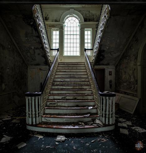 A beautiful white wooden staircase leading up to a landing with three large windows. The area is badly deteriorated with black mold creeping across the walls