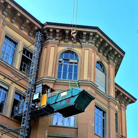 Outside picture of a building in classical style, with light scaffolding reaching up to the crowning. The scaffolding connects to a skip container, held up by a crane hook.