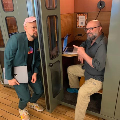 Jeff sits in a "phone booth" with his laptop open. He points at Ken who is positioned to peer through a window in the door of the phone booth. However, the door is open.  They're both looking at the camera