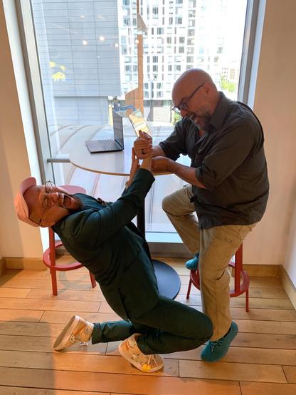 Ken playfully throws his head back with his eyes closed and mouth agape as he falls to his knees.  Jeff continues to hold Ken's hand in mock victory from their arm-wrestling match. His face is maniacal and joyful. He's still half-seated on the bar stool.